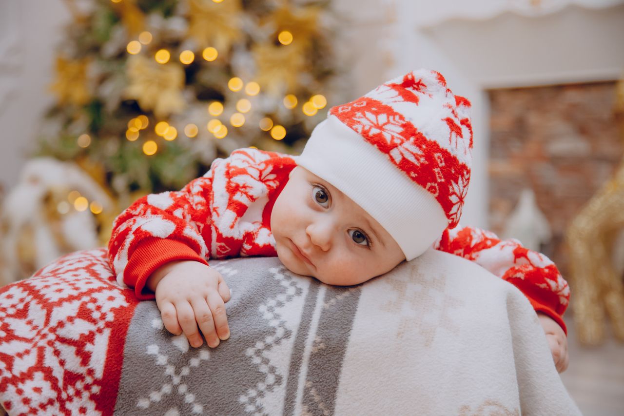 Charming baby in festive red attire resting beside a decorated Christmas tree indoors.